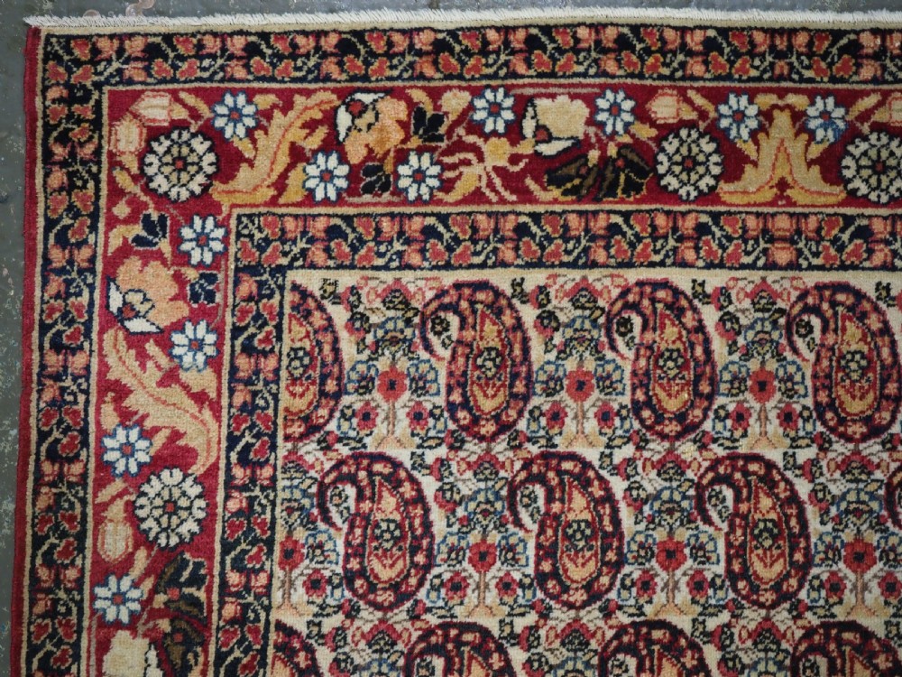 An antique Kirman rug featuring a repeat floral boteh pattern on a cream field enclosed by a detailed red and cream floral border. View VI