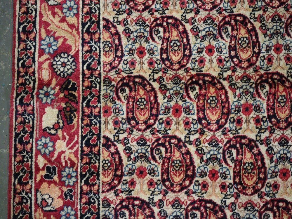 An antique Kirman rug featuring a repeat floral boteh pattern on a cream field enclosed by a detailed red and cream floral border. View VII