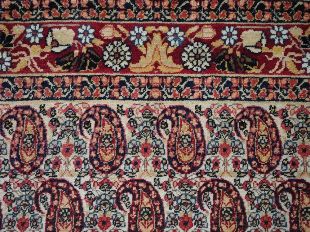 An antique Kirman rug featuring a repeat floral boteh pattern on a cream field enclosed by a detailed red and cream floral border. View VIII
