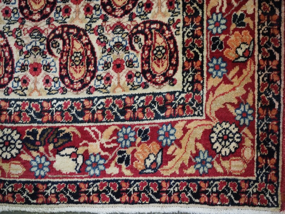 An antique Kirman rug featuring a repeat floral boteh pattern on a cream field enclosed by a detailed red and cream floral border. View X