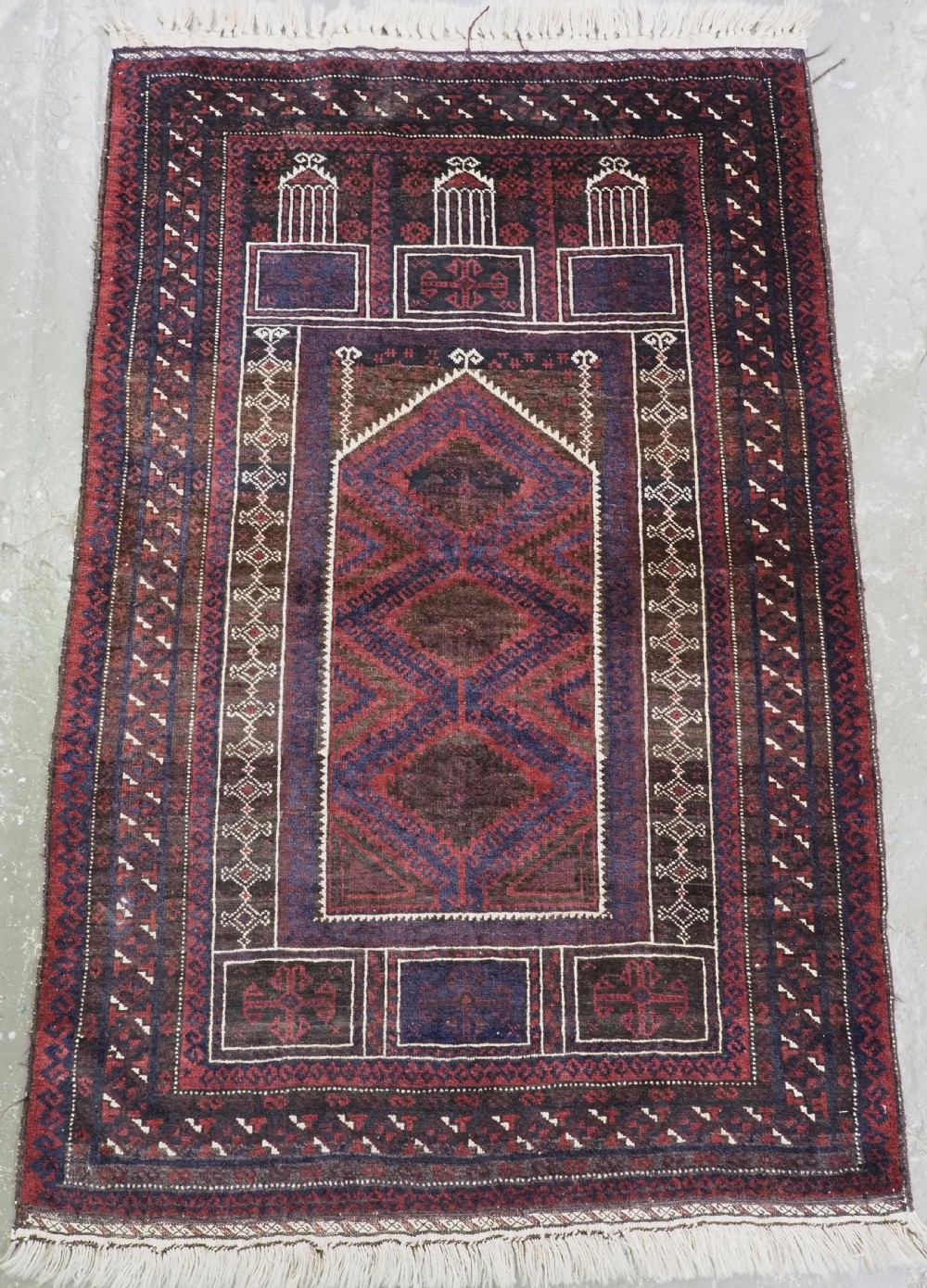 An antique rectangular Baluch wool rug featuring a central geometric Seh Mihrab prayer arch in deep red, indigo, and dark brown tones.