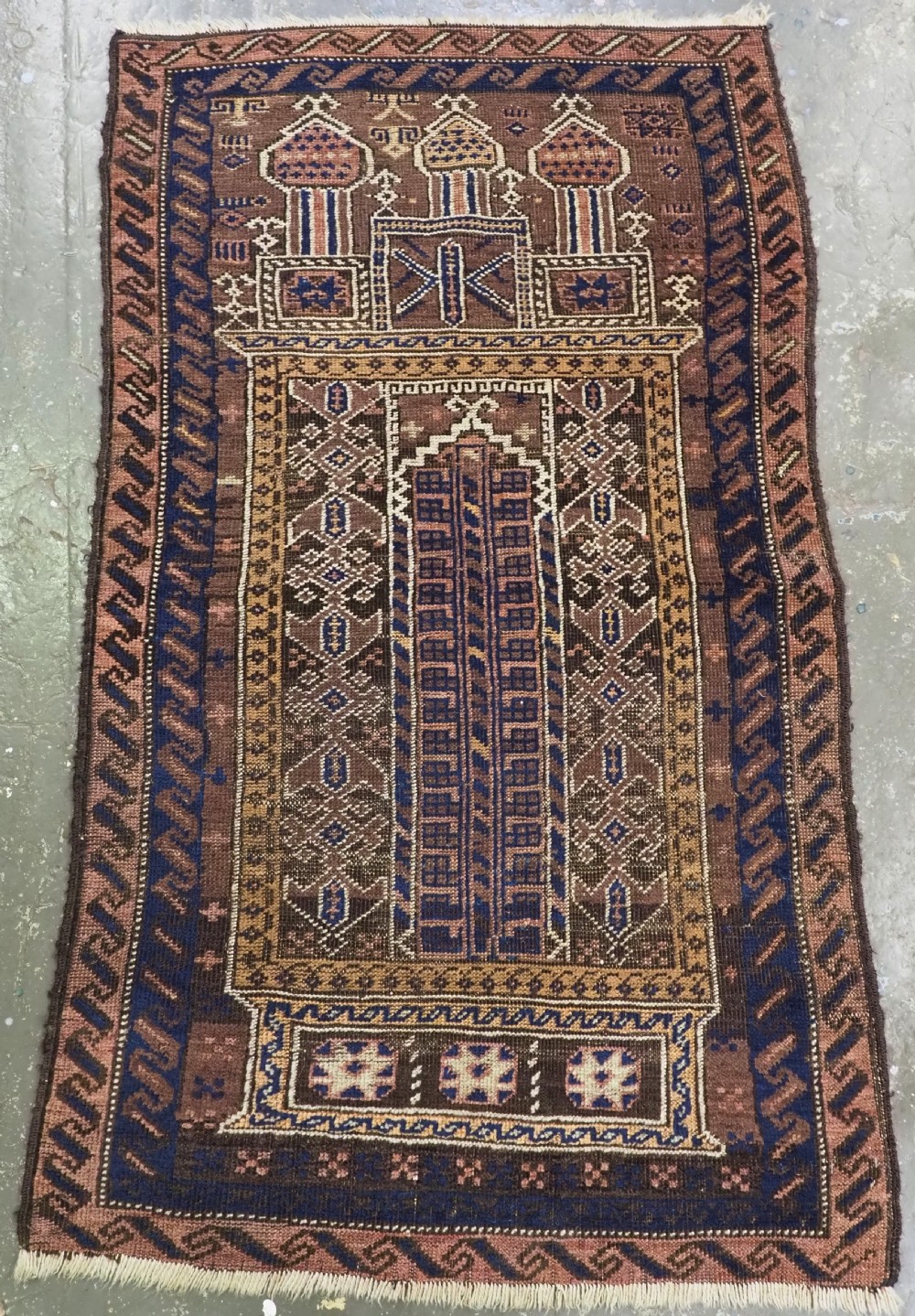 An antique Afghan Baluch handwoven wool prayer rug featuring geometric triple mihrab arches in shades of dark brown, navy blue, and terracotta.