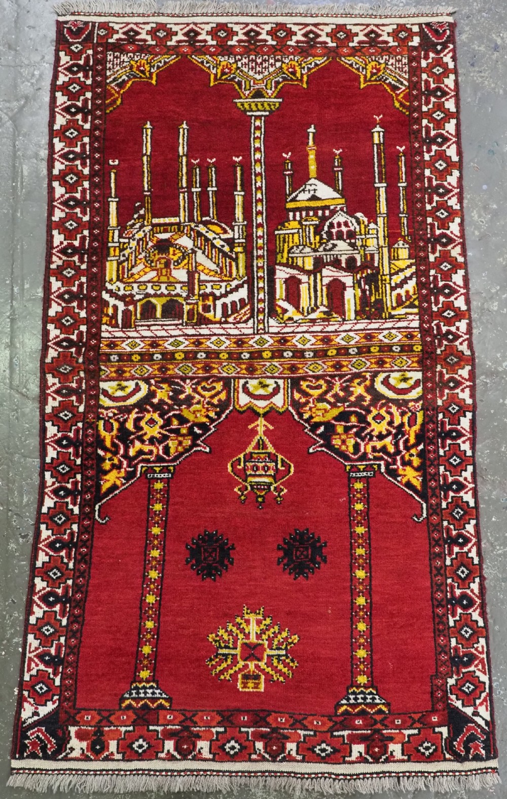 An antique hand-knotted wool prayer rug featuring architectural mosque motifs and geometric borders in deep red, yellow, cream, and black.