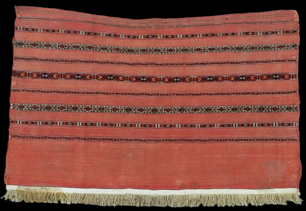 An antique Turkmen woven chuval face featuring horizontal bands of terracotta red and narrow geometric patterns in dark blue, black, and white with a fringed bottom edge.