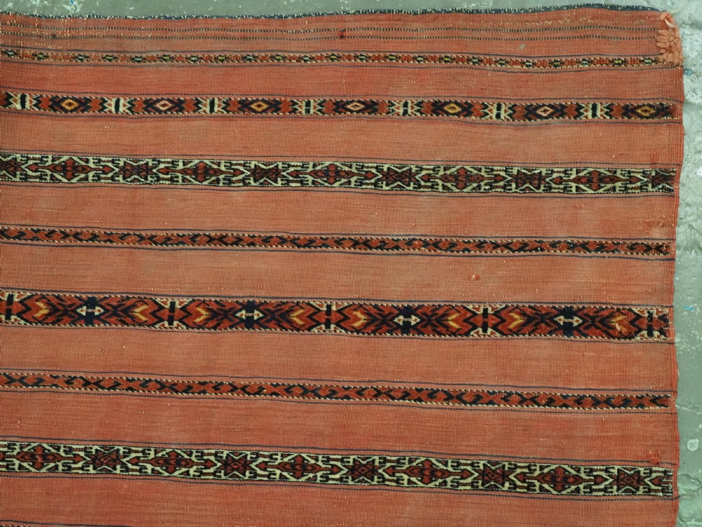An antique Turkmen woven chuval face featuring horizontal bands of terracotta red and narrow geometric patterns in dark blue, black, and white with a fringed bottom edge. View IV
