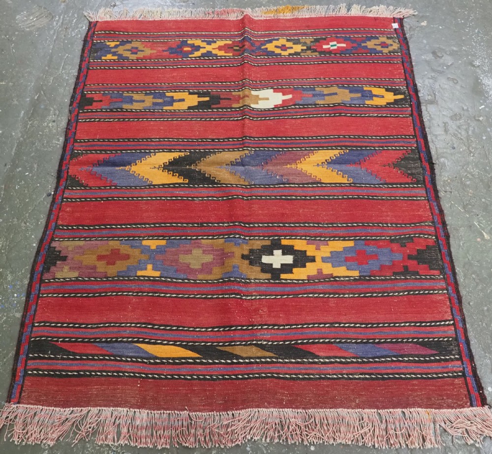 An antique flatweave Baluch kilim rug featuring horizontal red bands interspaced with colourful chevron and geometric motifs.