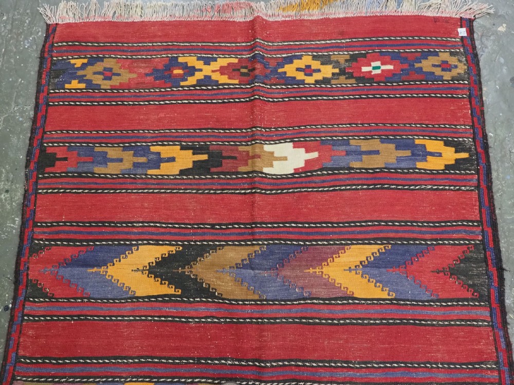 An antique flatweave Baluch kilim rug featuring horizontal red bands interspaced with colourful chevron and geometric motifs. View III