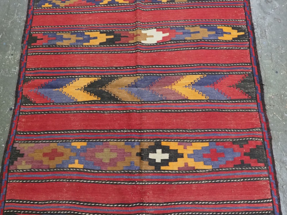 An antique flatweave Baluch kilim rug featuring horizontal red bands interspaced with colourful chevron and geometric motifs. View IV