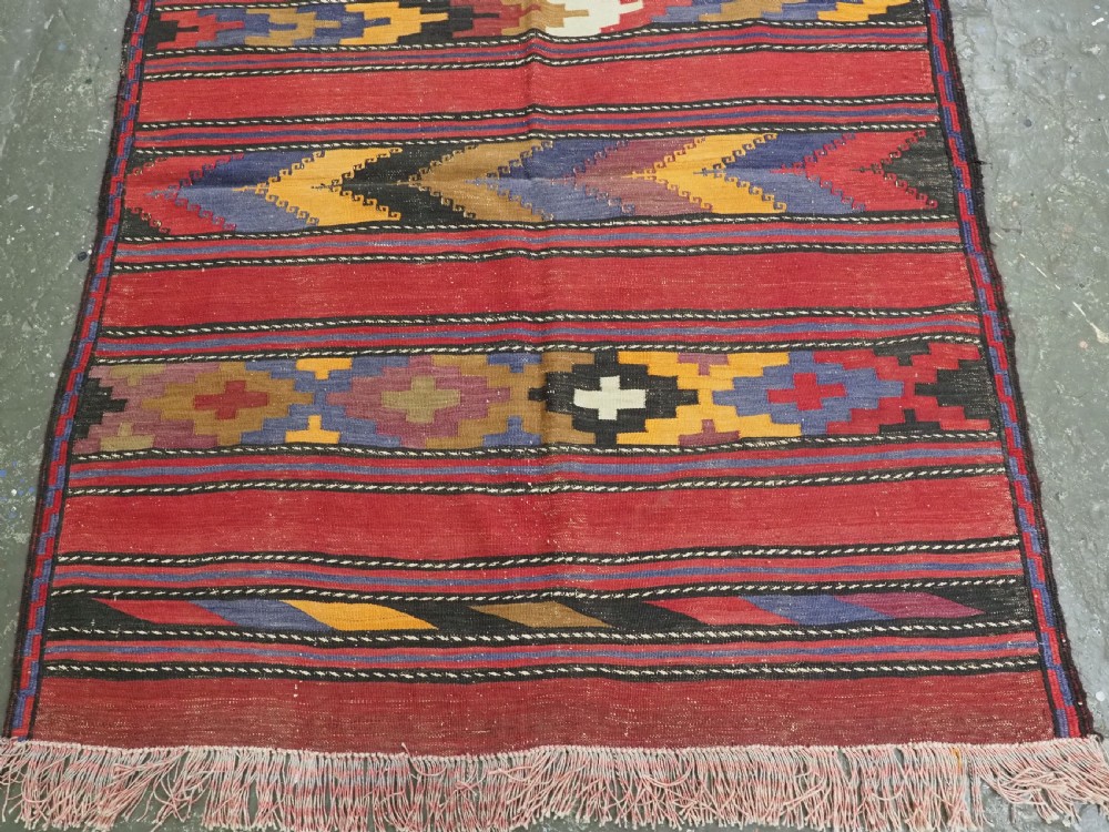 An antique flatweave Baluch kilim rug featuring horizontal red bands interspaced with colourful chevron and geometric motifs. View V