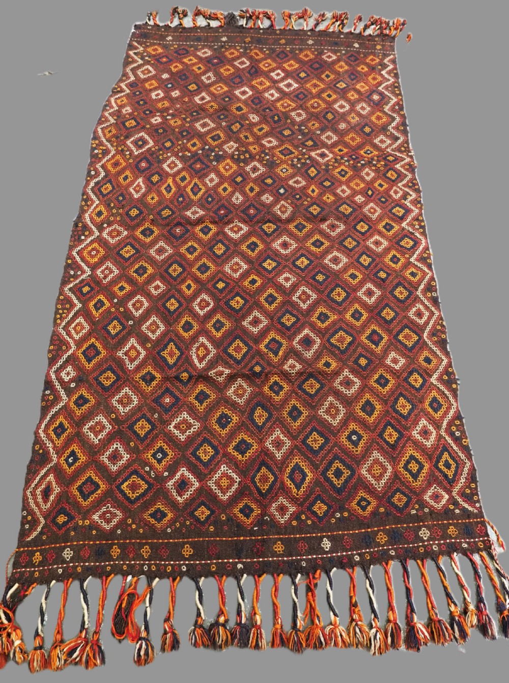 A hand-woven antique Afghan kilim textile featuring dark brown wool embroidered with a geometric diamond lattice pattern in rust, yellow, and cream, finished with long braided fringe tassels.