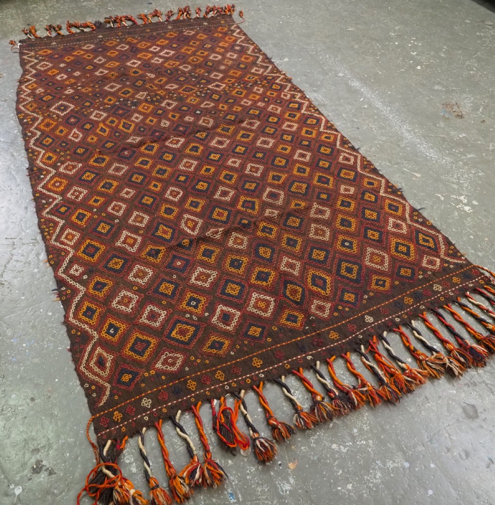 A hand-woven antique Afghan kilim textile featuring dark brown wool embroidered with a geometric diamond lattice pattern in rust, yellow, and cream, finished with long braided fringe tassels. View II