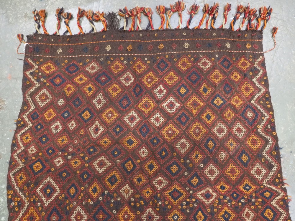 A hand-woven antique Afghan kilim textile featuring dark brown wool embroidered with a geometric diamond lattice pattern in rust, yellow, and cream, finished with long braided fringe tassels. View III