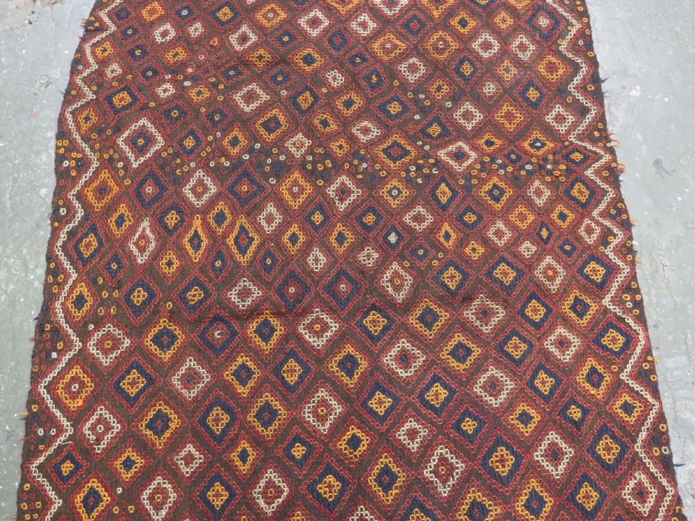 A hand-woven antique Afghan kilim textile featuring dark brown wool embroidered with a geometric diamond lattice pattern in rust, yellow, and cream, finished with long braided fringe tassels. View IV