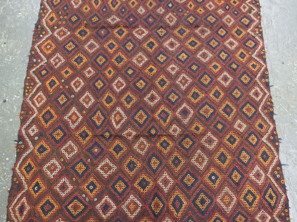 A hand-woven antique Afghan kilim textile featuring dark brown wool embroidered with a geometric diamond lattice pattern in rust, yellow, and cream, finished with long braided fringe tassels. View V