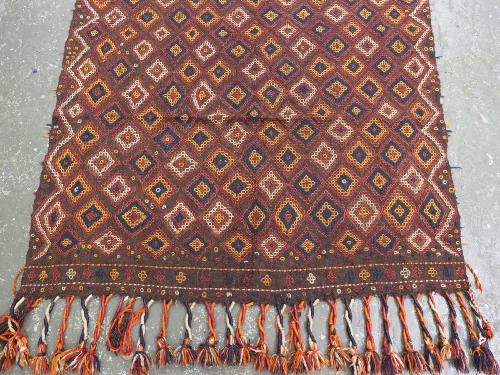 A hand-woven antique Afghan kilim textile featuring dark brown wool embroidered with a geometric diamond lattice pattern in rust, yellow, and cream, finished with long braided fringe tassels. View VI