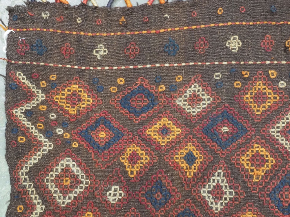A hand-woven antique Afghan kilim textile featuring dark brown wool embroidered with a geometric diamond lattice pattern in rust, yellow, and cream, finished with long braided fringe tassels. View VII