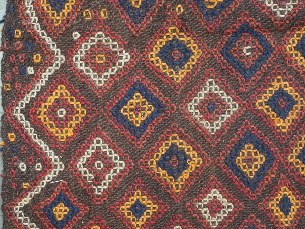 A hand-woven antique Afghan kilim textile featuring dark brown wool embroidered with a geometric diamond lattice pattern in rust, yellow, and cream, finished with long braided fringe tassels. View VIII