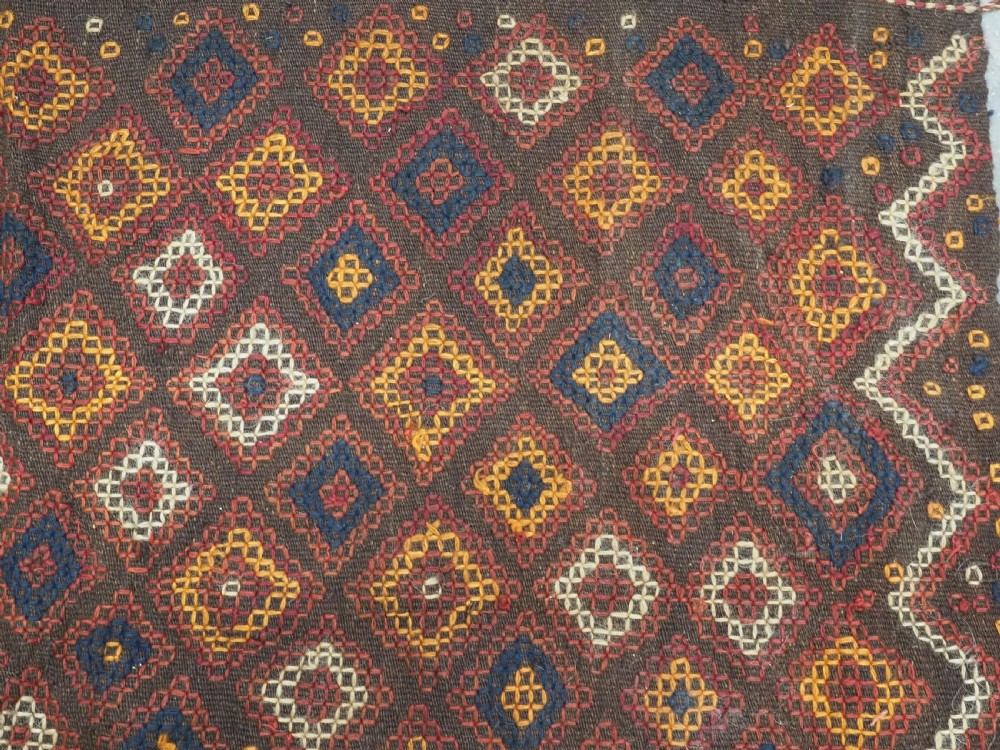 A hand-woven antique Afghan kilim textile featuring dark brown wool embroidered with a geometric diamond lattice pattern in rust, yellow, and cream, finished with long braided fringe tassels. View IX