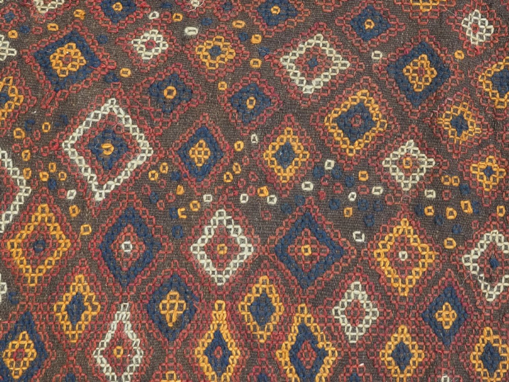 A hand-woven antique Afghan kilim textile featuring dark brown wool embroidered with a geometric diamond lattice pattern in rust, yellow, and cream, finished with long braided fringe tassels. View X