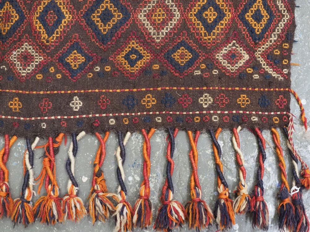 A hand-woven antique Afghan kilim textile featuring dark brown wool embroidered with a geometric diamond lattice pattern in rust, yellow, and cream, finished with long braided fringe tassels. View XIII
