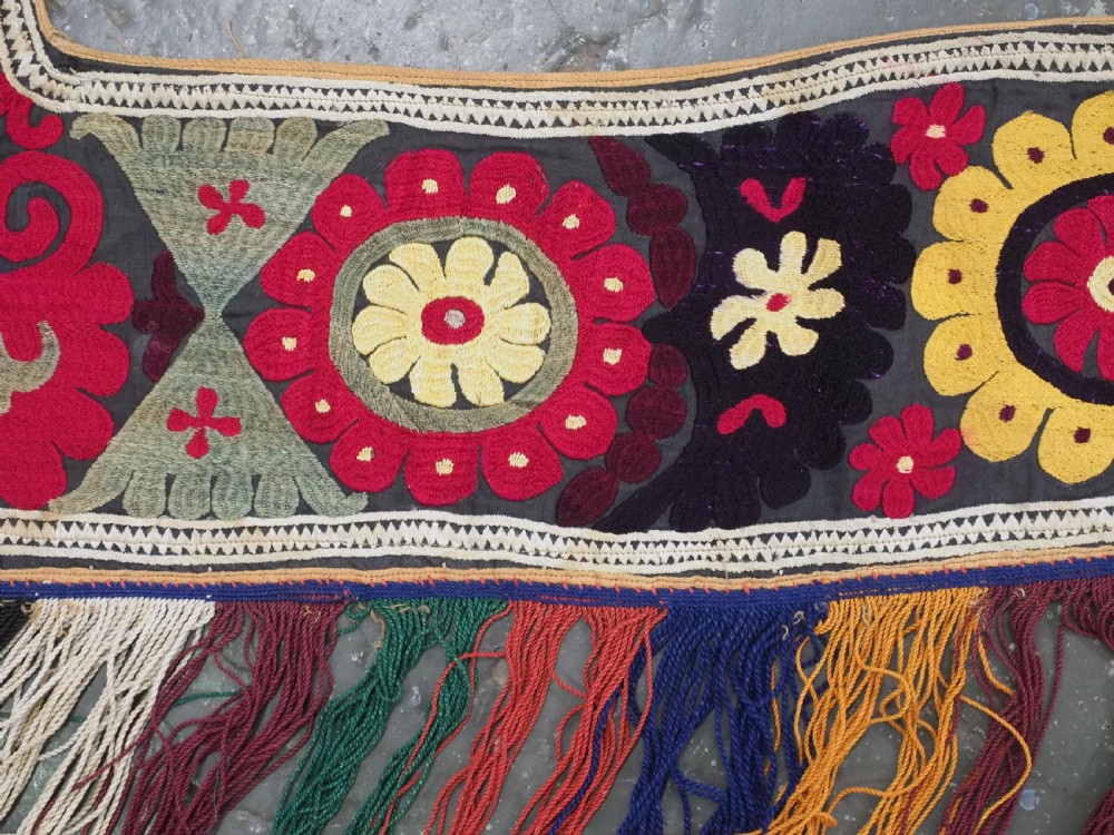 A V-shaped black cloth band decorated with vibrant red and yellow silk embroidered rosettes and finished with multi-coloured yarn fringe. View V