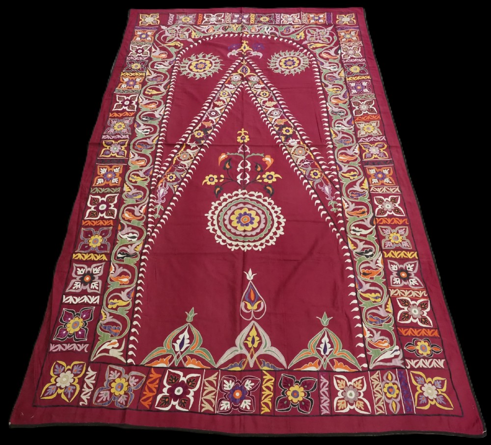 An antique rectangular Uzbek suzani featuring silk floral needlework embroidery on a deep red cotton ground with a pointed arch motif and floral borders.