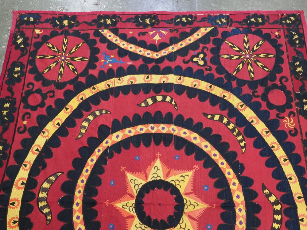 A rectangular crimson Uzbek suzani textile featuring heavy silk embroidery with a central star motif inside concentric circular borders. View II