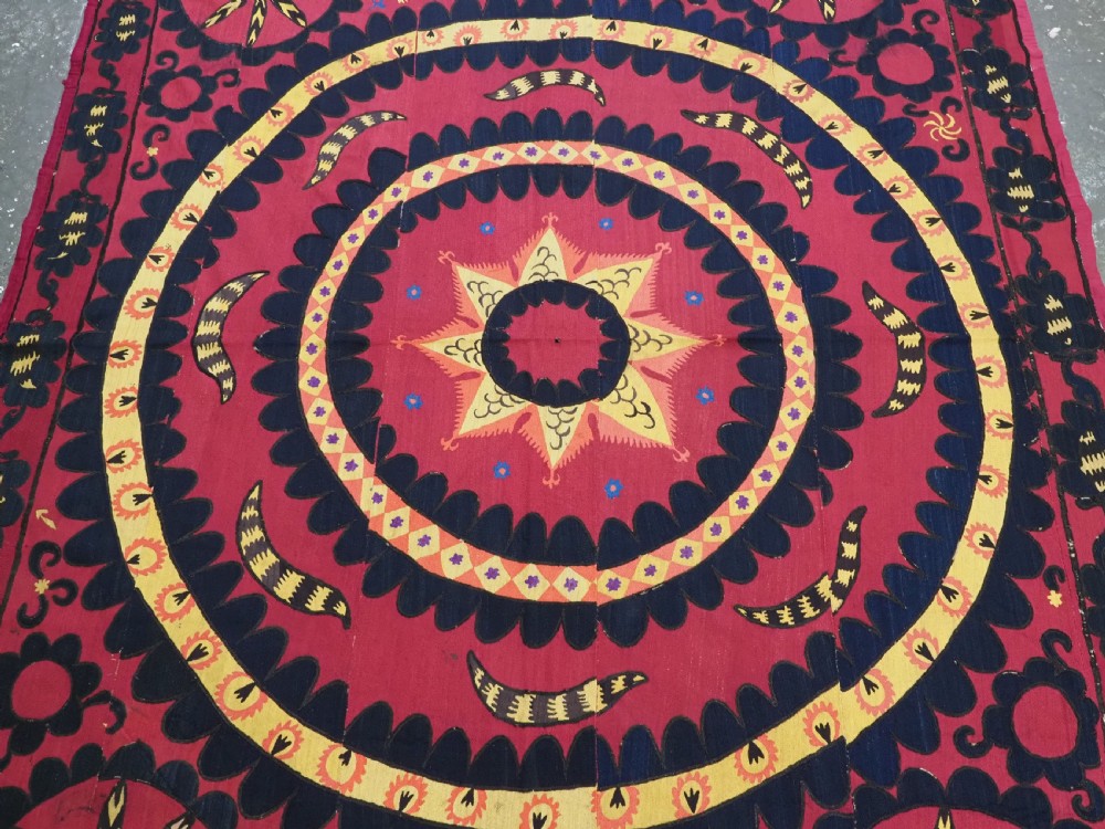 A rectangular crimson Uzbek suzani textile featuring heavy silk embroidery with a central star motif inside concentric circular borders. View III