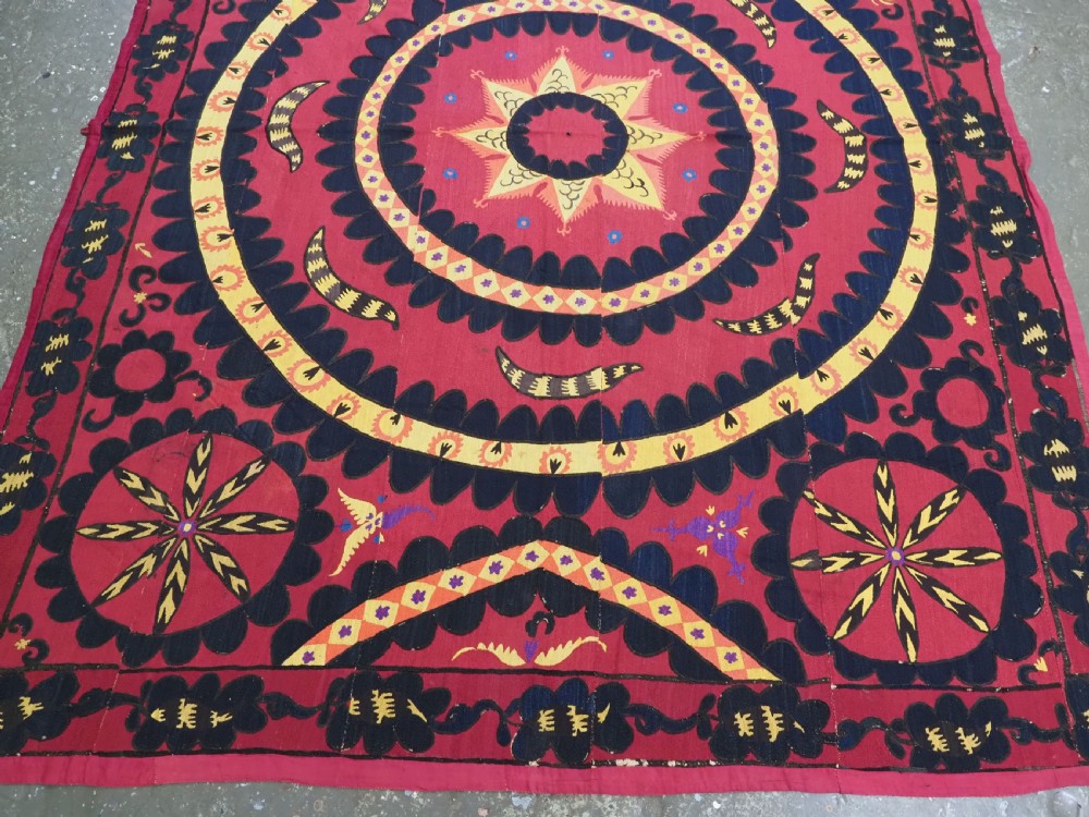 A rectangular crimson Uzbek suzani textile featuring heavy silk embroidery with a central star motif inside concentric circular borders. View IV