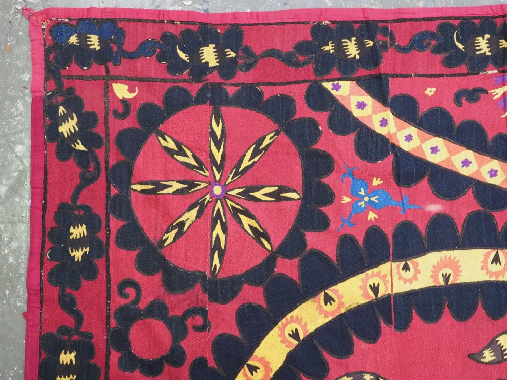 A rectangular crimson Uzbek suzani textile featuring heavy silk embroidery with a central star motif inside concentric circular borders. View V