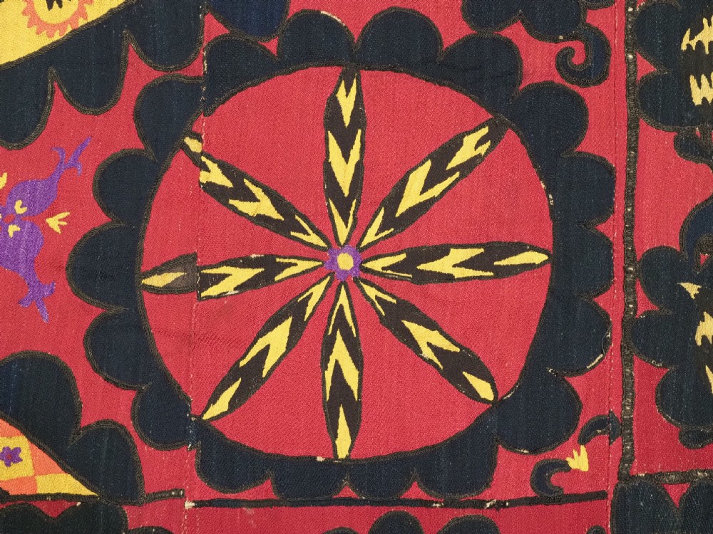 A rectangular crimson Uzbek suzani textile featuring heavy silk embroidery with a central star motif inside concentric circular borders. View X