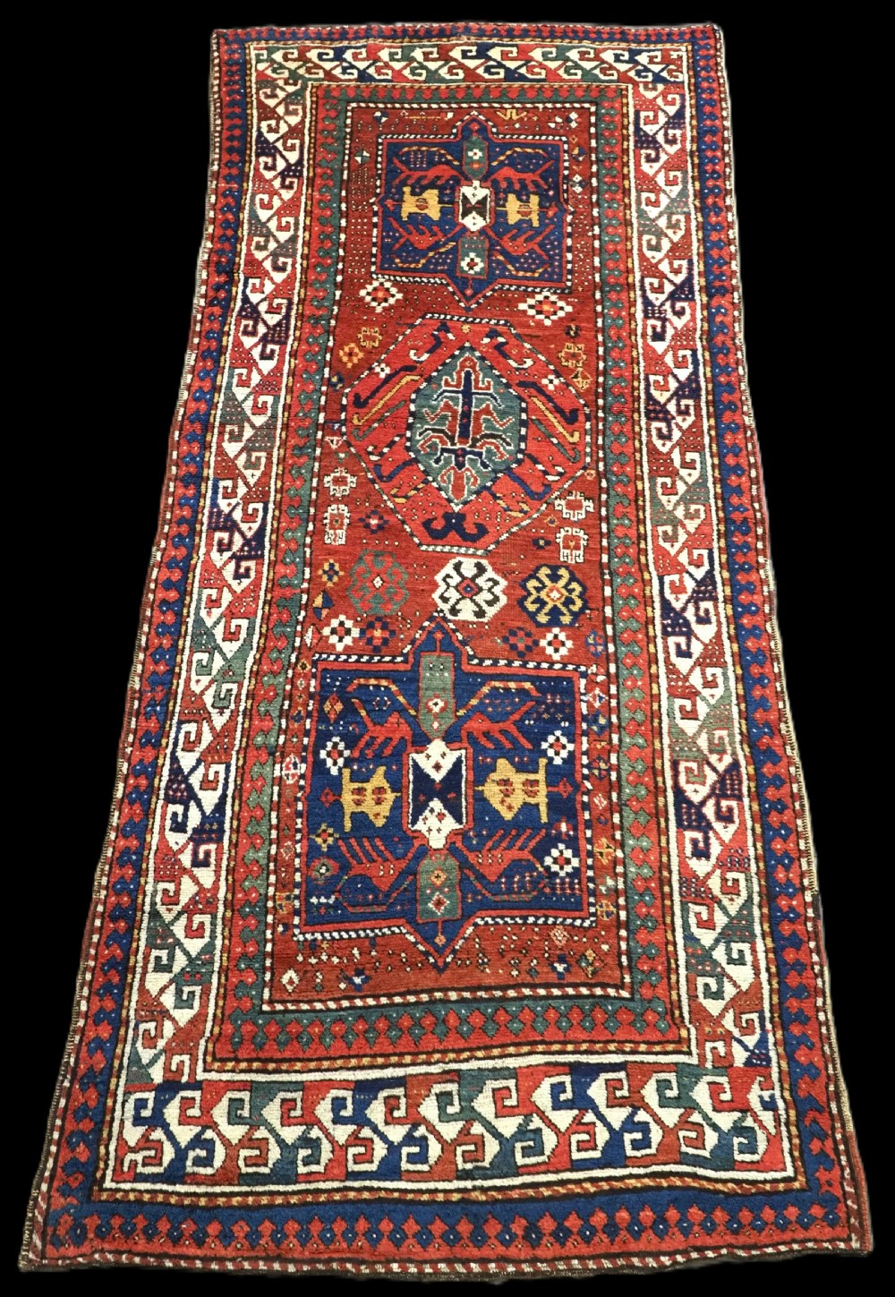 An antique Caucasian Kazak long rug featuring central geometric medallions in red, navy blue, and green with detailed border patterns.