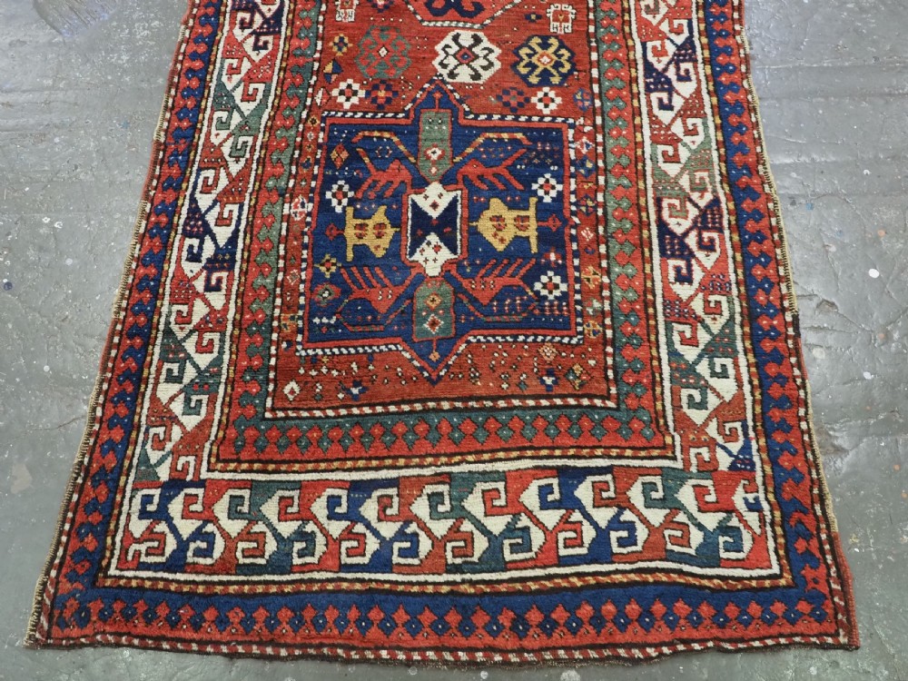An antique Caucasian Kazak long rug featuring central geometric medallions in red, navy blue, and green with detailed border patterns. View IV