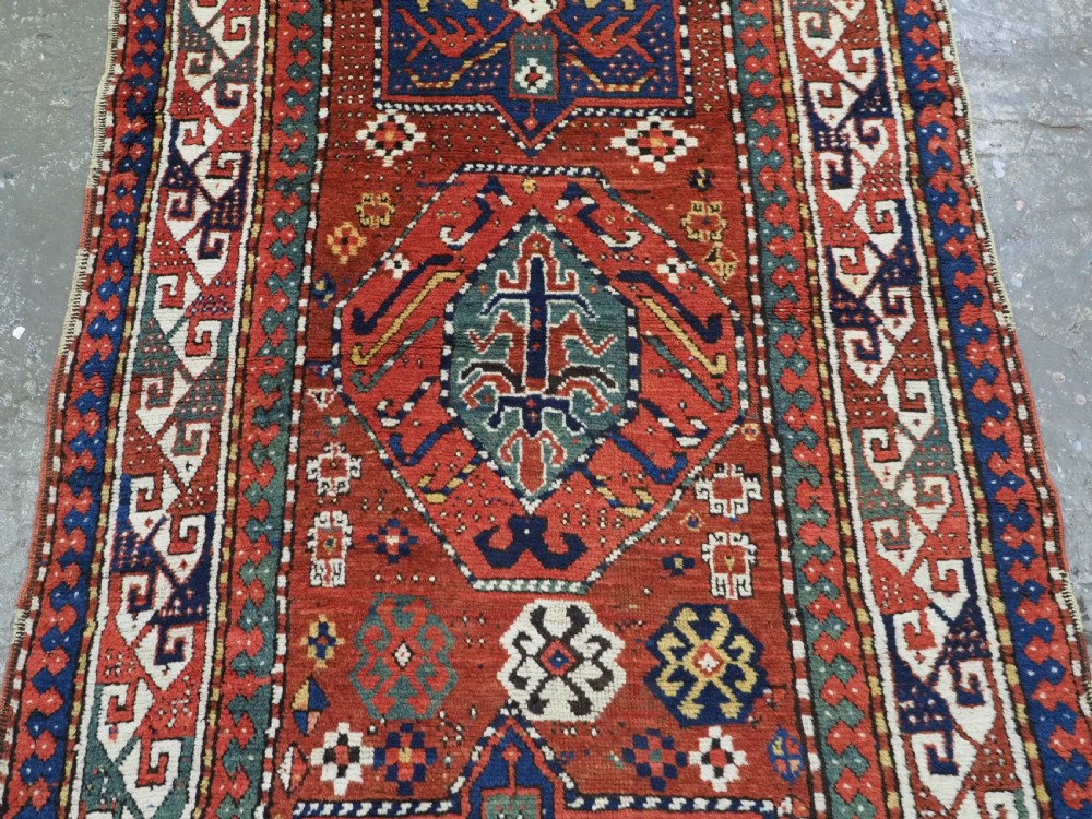 An antique Caucasian Kazak long rug featuring central geometric medallions in red, navy blue, and green with detailed border patterns. View V