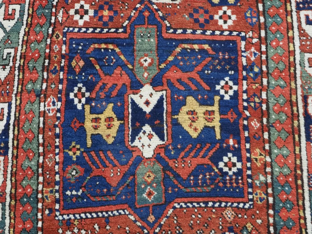 An antique Caucasian Kazak long rug featuring central geometric medallions in red, navy blue, and green with detailed border patterns. View X