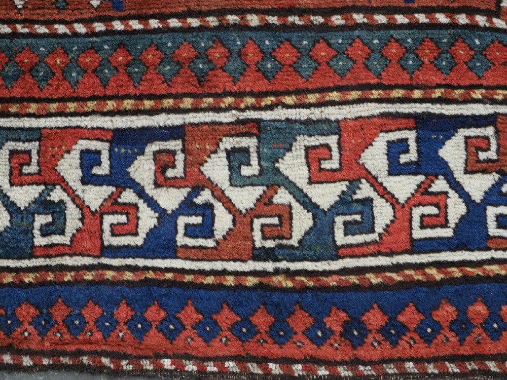 An antique Caucasian Kazak long rug featuring central geometric medallions in red, navy blue, and green with detailed border patterns. View XII