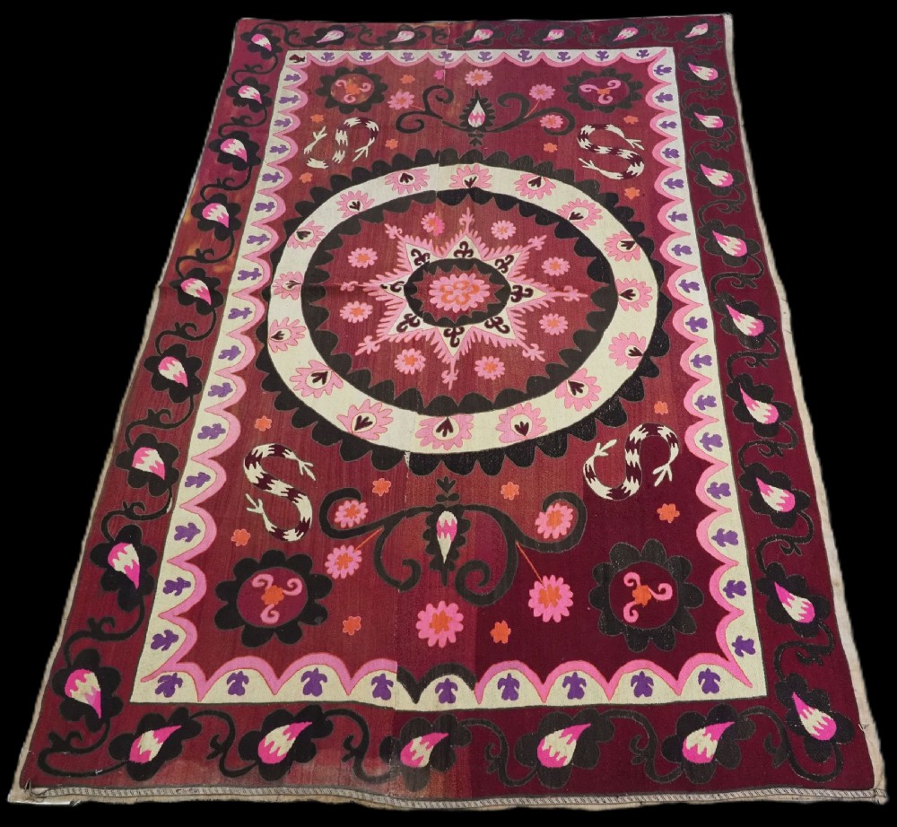 A rectangular hand-embroidered textile featuring a large central circular starburst medallion surrounded by floral motifs in shades of pink, cream, orange, and burgundy.