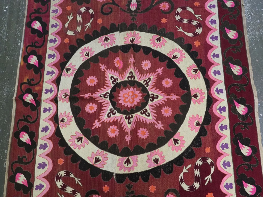 A rectangular hand-embroidered textile featuring a large central circular starburst medallion surrounded by floral motifs in shades of pink, cream, orange, and burgundy. View III