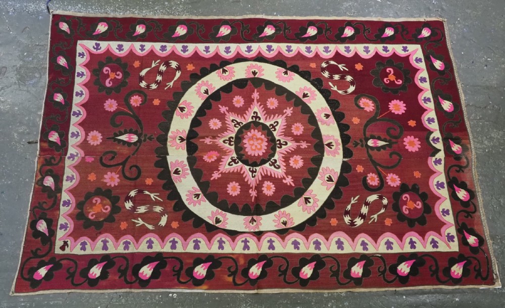 A rectangular hand-embroidered textile featuring a large central circular starburst medallion surrounded by floral motifs in shades of pink, cream, orange, and burgundy. View IV