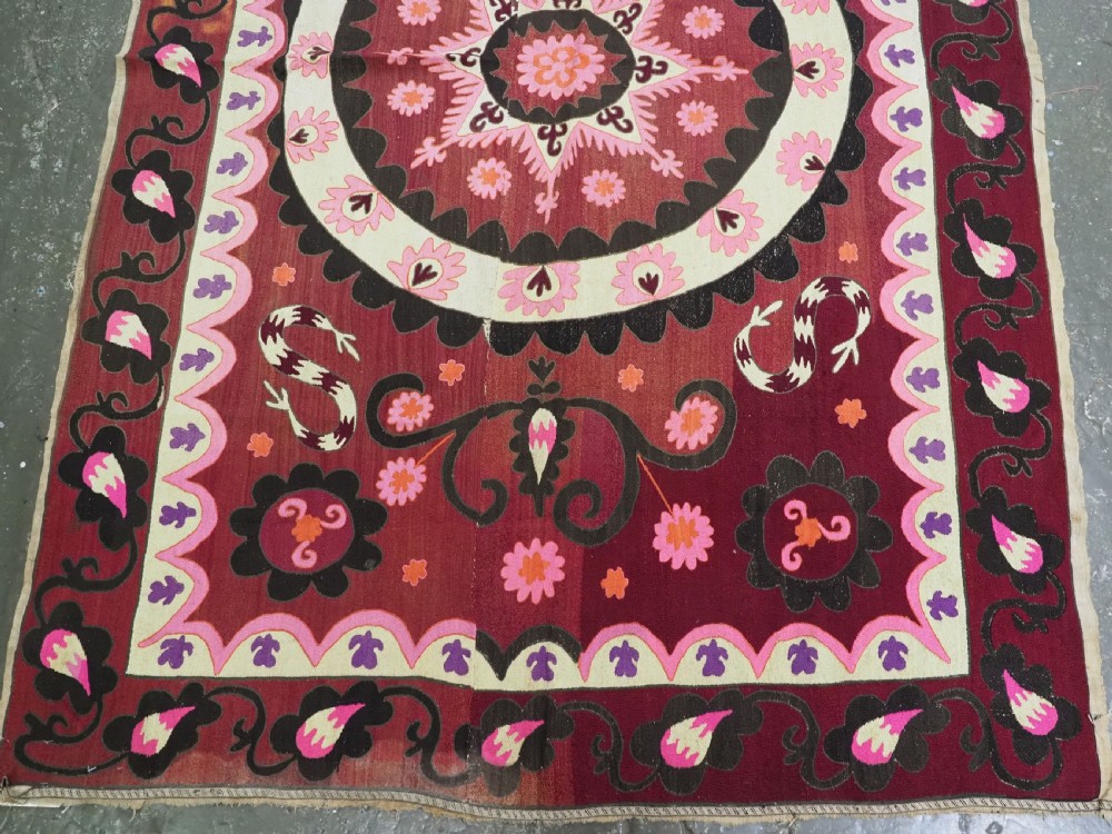 A rectangular hand-embroidered textile featuring a large central circular starburst medallion surrounded by floral motifs in shades of pink, cream, orange, and burgundy. View V