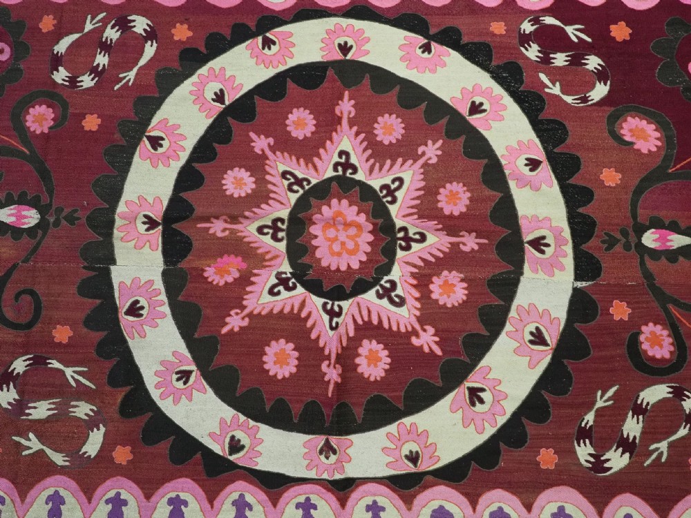 A rectangular hand-embroidered textile featuring a large central circular starburst medallion surrounded by floral motifs in shades of pink, cream, orange, and burgundy. View VII