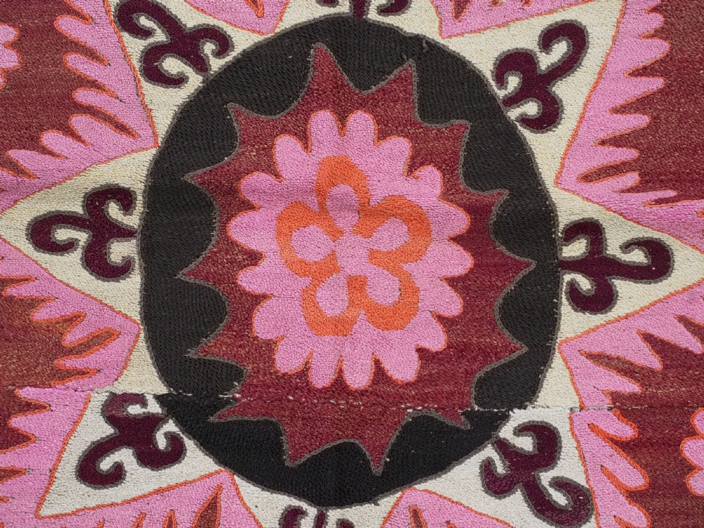 A rectangular hand-embroidered textile featuring a large central circular starburst medallion surrounded by floral motifs in shades of pink, cream, orange, and burgundy. View XII