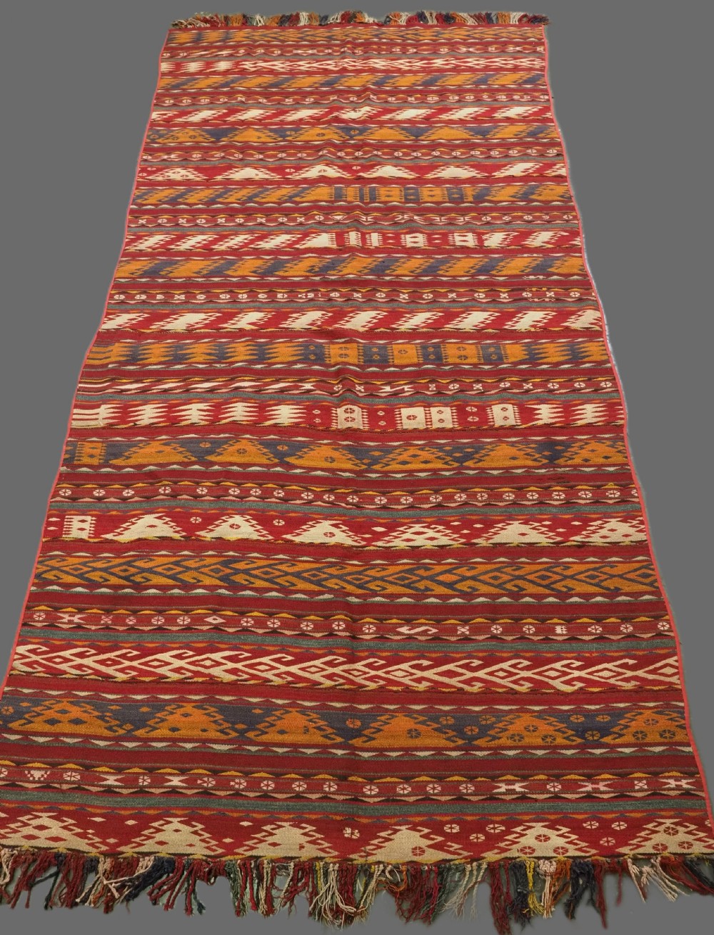 An antique Central Asian Uzbek Gajeri flatweave kilim rug featuring horizontal bands of geometric motifs woven in red, ochre, navy, and cream wool with fringed ends.