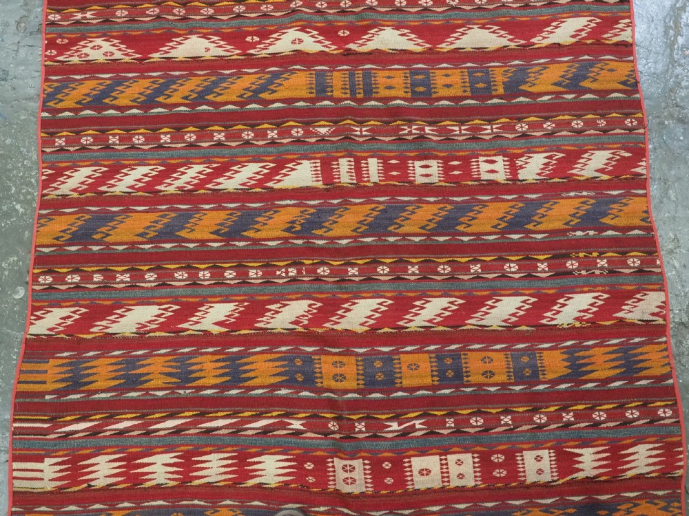 An antique Central Asian Uzbek Gajeri flatweave kilim rug featuring horizontal bands of geometric motifs woven in red, ochre, navy, and cream wool with fringed ends. View IV