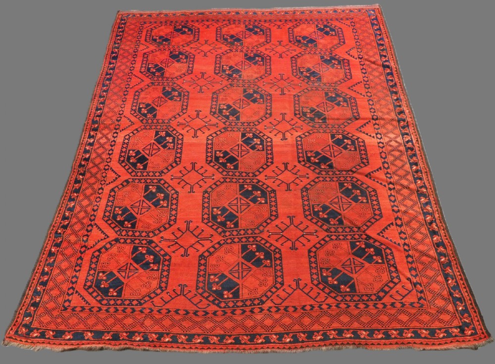 An antique handwoven Ersari Turkmen carpet featuring repeating octagonal guls in dark navy and red set upon a vibrant terracotta red background with geometric borders.