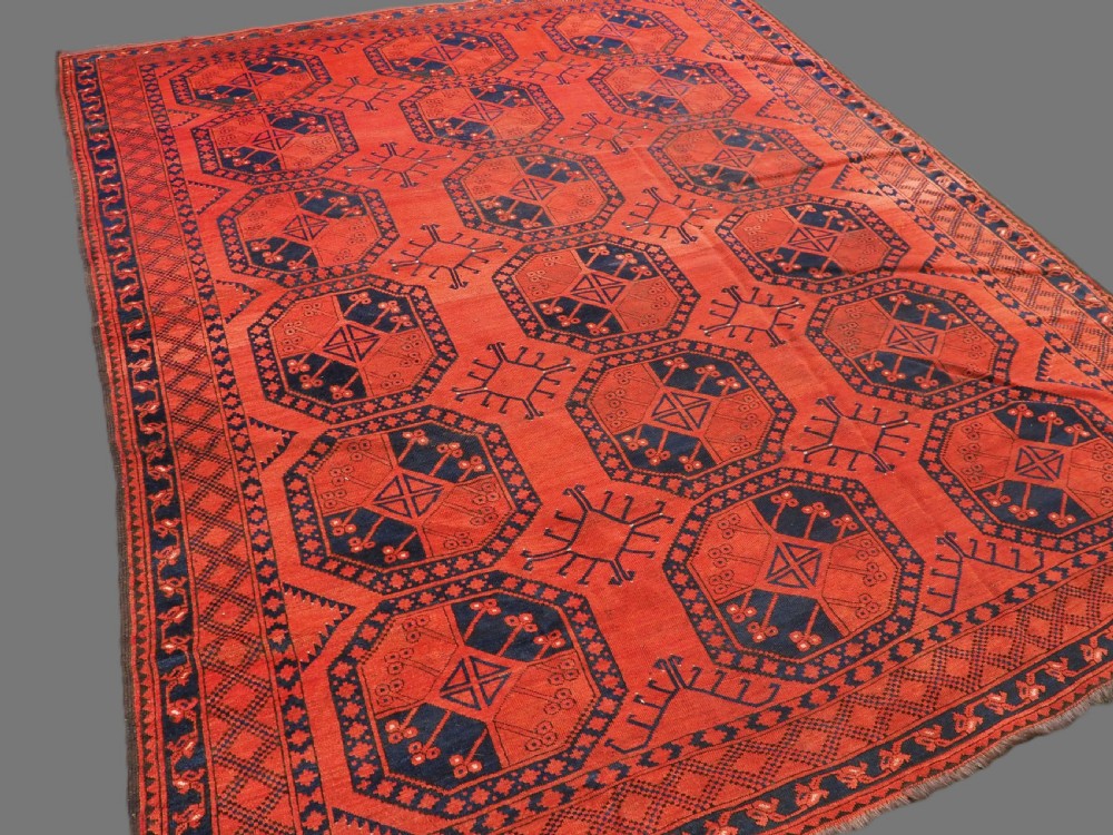 An antique handwoven Ersari Turkmen carpet featuring repeating octagonal guls in dark navy and red set upon a vibrant terracotta red background with geometric borders. View II
