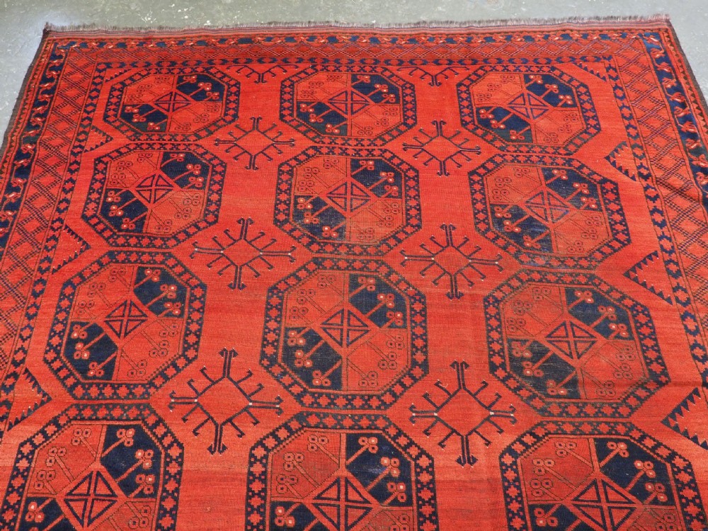 An antique handwoven Ersari Turkmen carpet featuring repeating octagonal guls in dark navy and red set upon a vibrant terracotta red background with geometric borders. View III
