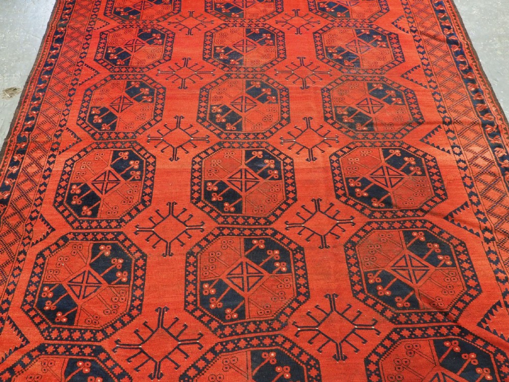 An antique handwoven Ersari Turkmen carpet featuring repeating octagonal guls in dark navy and red set upon a vibrant terracotta red background with geometric borders. View IV