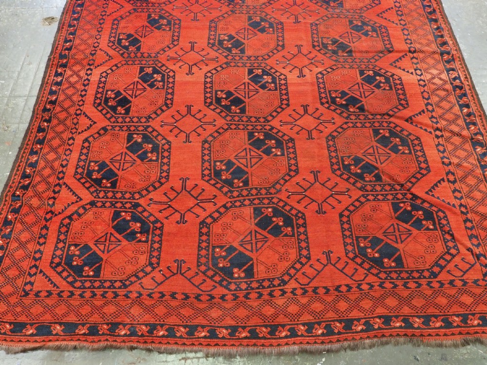An antique handwoven Ersari Turkmen carpet featuring repeating octagonal guls in dark navy and red set upon a vibrant terracotta red background with geometric borders. View V