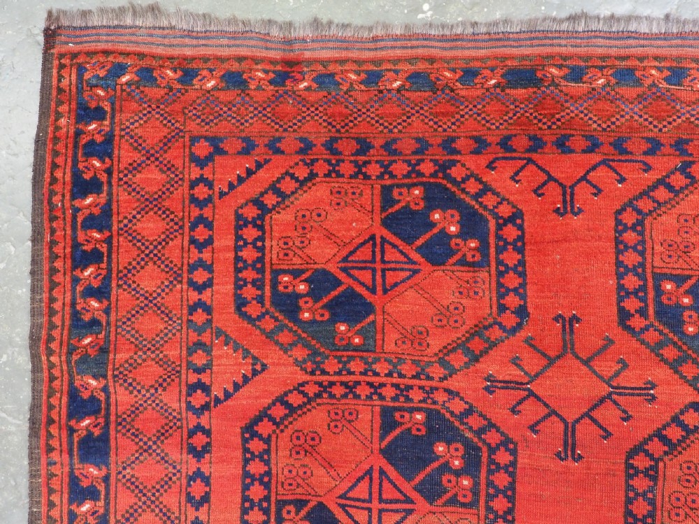 An antique handwoven Ersari Turkmen carpet featuring repeating octagonal guls in dark navy and red set upon a vibrant terracotta red background with geometric borders. View VI
